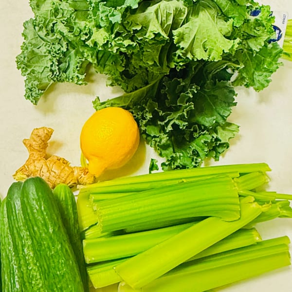 Fresh green kale, celery, cucumbers, a lemon, and ginger root on a light surface.